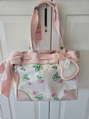 Juicy Couture daydreamer gelato rosa körsbär - Söt handväska från Juicy Couture i ljusrosa och vitt med gröna och rosa körsbärsmotiv. Väskan har rosa detaljer, ett hjärtformat nyckelband och en mjuk scarf knuten på handtaget. Perfekt för dig som gillar gulliga accessoarer och Y2K-stil.