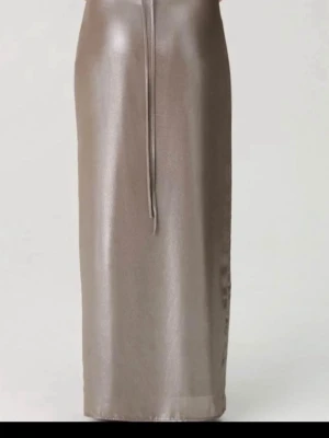 Lång metallic kjol med slits - Snygg långkjol i skimrande silverfärg med hög slits framtill. Kjolen har en tight passform och är tillverkad i ett glansigt satinliknande material. Perfekt för dig som vill sticka ut med en trendig och elegant look.