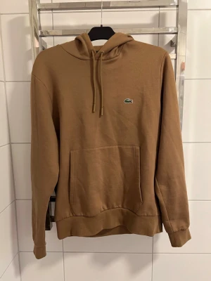 Lacoste Hoodie  - Snygg brun hoodie från Lacoste med klassisk krokodillogga på bröstet. Hoodien har huva med dragsko och en stor magficka framtill. Perfekt för en avslappnad och trendig stil.