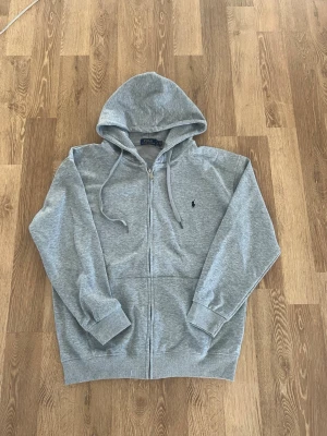 Grå zip-hoodie från Polo Ralph Lauren - Snygg grå hoodie med dragkedja från Polo Ralph Lauren. Klassisk design med huva och dragsnören, två fickor fram och broderad logga på bröstet. Tillverkad i mjuk bomullsblandning som är skön att ha på sig.