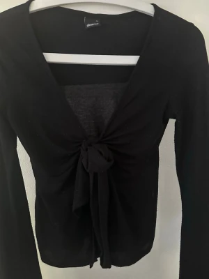 Svart långärmad knyt-topp från Gina Tricot - Svart långärmad topp från Gina Tricot med djup v-ringning och knyt framtill. Tunn och lätt i materialet, perfekt för lager-på-lager. Snygg och enkel att styla till olika outfits.