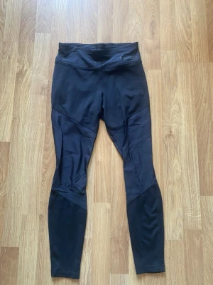 Svarta leggings från Stronger - Svarta leggings från Stronger med hög midja och diskret broderad logga på linningen. Tillverkade i stretchigt funktionsmaterial som sitter tight och är perfekta för träning eller chill. Snygga sömdetaljer längs benen för extra stilpoäng.