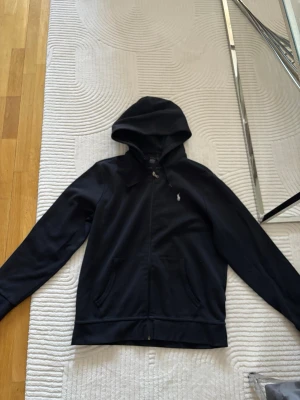 Svart hoodie från Polo Ralph Lauren - Svart hoodie med dragkedja från Polo Ralph Lauren. Klassisk design med huva och dragsko, två fickor framtill och den ikoniska broderade loggan på bröstet. Perfekt för en chill och stilren look. Materialet känns mjukt och skönt mot huden.   Snöret vid luvan är lite trasigt men går att fixa 