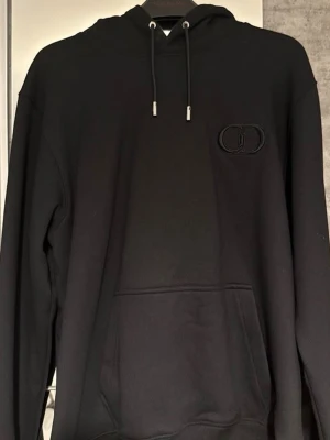 Svart hoodie från CD med logga - Svart hoodie från CD med broderad CD-logga på bröstet. Klassisk modell med huva, dragsnören och stor magficka. Tillverkad i mjukt material som känns skönt mot huden. Perfekt för en stilren och avslappnad look.