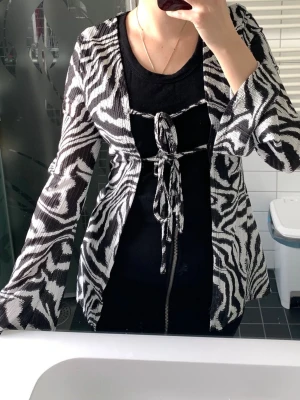 Zebramönstrad svartvit kofta med snörning - Cool zebramönstrad kofta i svart och vitt med långa ärmar och snörningsdetaljer framtill. Koftan har en loose passform och är perfekt att slänga över en enkel topp för att ge outfiten ett lyft. Materialet känns lätt och bekvämt. 