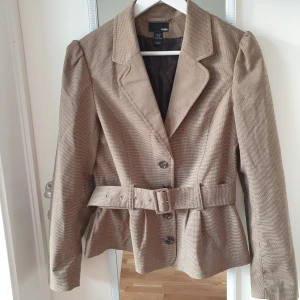 Beige kavaj - Snygg beige kavaj från H&M med ett diskret rutigt mönster. Kavajen har långa ärmar och knäpps med knappar framtill. Den har också ett bälte i midjan för en mer definierad passform. Perfekt för en stilren look. 