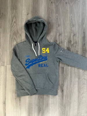 Grå Superdry hoodie med tryck - Snygg grå hoodie från Superdry med blått och gult tryck på bröstet där det står 'SuperDry REAL' och siffran 54🤩 storlek S, inga fel😇 hör av er vid funderingar samt snabbaffär🙌🏻