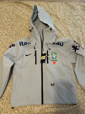 Nike Brasil windbreaker med huva - Ljusgrå windbreaker från Nike med Brasil-logga och sponsormärken på bröst och ärmar. Jackan har hel dragkedja, två fickor framtill och justerbar huva. Perfekt för dig som gillar sportig stil och vill visa stöd för Brasilien.