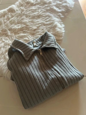 Gråblå ribbad ziptröja med krage - Snygg gråblå ribbstickad tröja med dragkedja framtill och bred krage. Tröjan har långa ärmar och är figurnära i modellen. Perfekt för lager-på-lager och enkel att styla till olika outfits.
