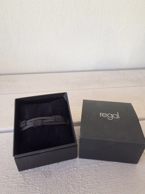 Svart läderarmband från Regal - Snyggt svart armband från Regal i läder med flätade detaljer och tvinnade snören. Armbandet har en stilren och modern design som passar perfekt till många olika outfits. Levereras i original ask.