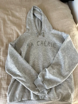Grå hoodie med texten MA CHÉRIE - Snygg grå hoodie med huva och dragsko, dekorerad med texten 'MA CHÉRIE' i glittriga stenar på bröstet. Mjuk och skön bomullsblandning, perfekt för en chill och avslappnad stil.