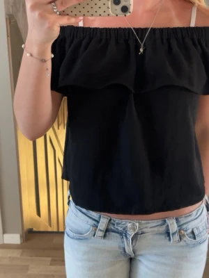 Svart offshoulder blus med volang - Stilren svart offshoulder blus med bred volang runt axlarna. Blusen har en rak och croppad passform och är tillverkad i ett mjukt, lätt tyg som känns skönt mot huden. Perfekt för sommarens varma dagar och enkel att matcha.