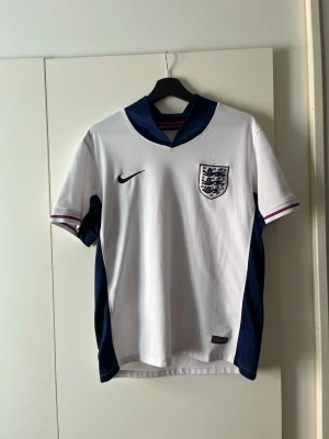 England Nike fotbollströja vit/blå - Snygg England fotbollströja från Nike i vitt med mörkblå detaljer längs sidorna och kragen. Tröjan har korta ärmar, broderad England-logga på bröstet och Nike-logga. Tillverkad i lätt och ventilerande material, perfekt för träning eller match.