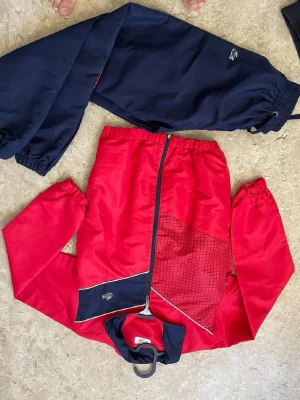 Rött och marinblått träningsset - Snyggt träningsset med jacka och byxor i rött och marinblått. Jackan har dragkedja, rutigt mönster på ena sidan och elastiska muddar. Byxorna är helblå med resår i midja och benslut. Perfekt för sportiga dagar eller chill.