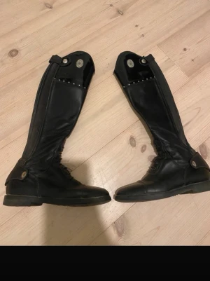 Romitelli ridstövlar - Säljer ett par svarta romitelli ridstövlar i äkta läder med högt skaft och dekorativa nitar upptill. Klassisk design med snörning vid vristen och knappar vid skaftets kant. Ena kedjan är trasig, bild finns. Även lite trasig högst upp på inner sidan men inget som syns. Kedjan går och laga lätt för någon hundralapp!