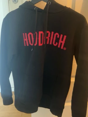 Svart hoodie från Hoodrich med tryck - Svart hoodie från Hoodrich med stor röd logga på bröstet. Klassisk modell med luva.