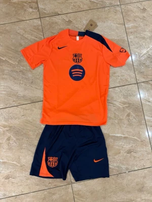Barça träningsset Nike orange/marin - Snyggt träningsset från Nike med FC Barcelona-logga. Tröjan är orange med marinblå detaljer och tryck, inklusive Spotify-logga och klubbmärke. Shortsen är marinblå med orange detaljer och klubbmärke. Perfekt för fotbollsträning eller sportiga dagar.