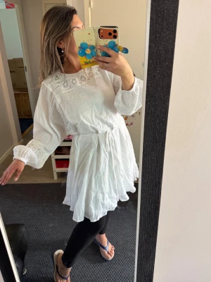 Vit kortklänning med spetsdetaljer - Säljer en vit kortklänning med långa ärmar och vacker spets runt halsen och ärmsluten. Klänningen har en omlott-effekt med knytband i midjan och är gjord i ett lätt bomullsmaterial som känns luftigt och skönt.Helt ny med prislapp 