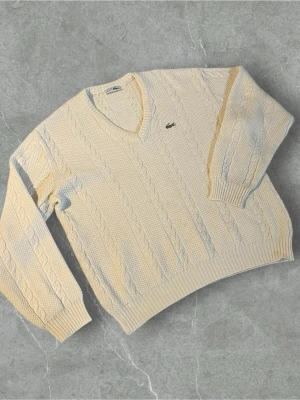 Krämvit kabelstickad tröja Lacoste | L - Snygg krämvit kabelstickad tröja från Lacoste med klassisk v-ringning och broderad krokodil-logga på bröstet. Tröjan har ribbade muddar och är tillverkad i mjukt stickat material. Perfekt för dig som gillar preppy stil och vill ha något bekvämt.