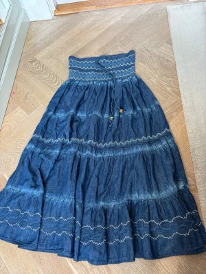 Blå långkjol med broderade detaljer - Säljer en blå långkjol med smockad midja och dekorativa broderade sömmar i vitt. Kjolen har volanger och är tillverkad i ett lätt bomullsmaterial som ger en skön och luftig känsla. Perfekt för dig som gillar boho-stilen och vill ha något unikt i garderoben. Skriv för fler bilder. Pris går att diskutera!🥰