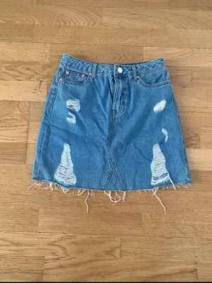 Blå jeanskjol med slitningar - Säljer en blå jeanskjol från Levi's med rå kant och tydliga slitningar framtill. Kjolen har klassisk femficksdesign, knapp och dragkedja samt fransig nederkant för en trendig look. Perfekt för dig som gillar en avslappnad och edgy stil.