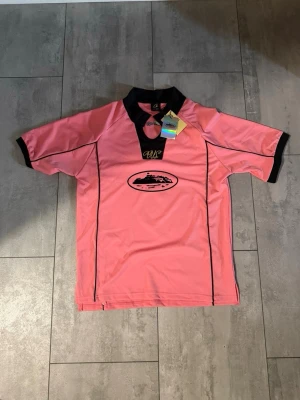 Cortiez jersey  - Snygg rosa sporttröja från Corte med svart krage och svarta detaljer vid ärmslut och längs sidorna. Tröjan har korta ärmar och ett tryckt motiv på bröstet samt broderad logga. Perfekt för träning eller lagidrott. Aldrig använd 