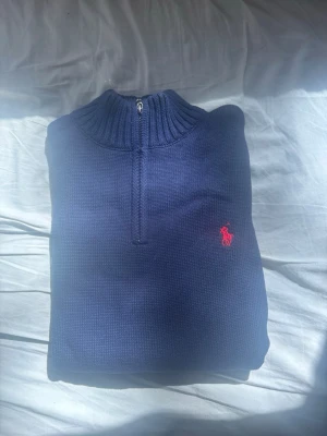 Blå stickad qzip Polo Ralph Lauren - Snygg blå stickad tröja från Polo Ralph Lauren med qzip och ribbad krage. Tröjan har det klassiska röda Polo-logot broderat på bröstet och är tillverkad i mjukt material som passar perfekt till lager-på-lager. Perfekt för dig som gillar stilrena plagg. Helt ny med tag kvar! Hör av er vid eventuella frågor