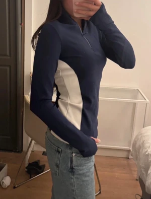 Marinblå långärmad topp med half zip - Säljer en mörkblå långärmad sporttröja med vita paneler på sidorna och halv dragkedja framtill. Tröjan har en kroppsnära passform och är perfekt för träning eller aktiva dagar. Materialet är troligtvis syntetiskt och designen är stilren och modern. Aldrig använt!! Helt ny 