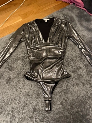 Metallic silver body med v-ringning - Snygg body i metallic silver med lång ärm och djup v-ringning. Kroppsnära passform och glansigt material som ger en cool look. Perfekt att styla med jeans eller kjol för en trendig outfit. Skulle även säga att den är lite tight i passform (den är storlek S men passar lite som en XS)