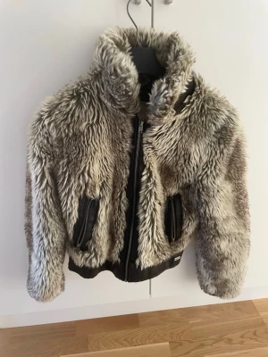 Pälsjacka från Crafted - Säljer en fluffig grå pälsjacka från Crafted med dragkedja framtill och två fickor med svarta detaljer. Jackan har hög krage och är tillverkad i fuskpäls