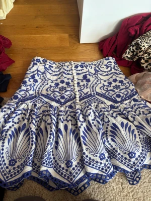 Vit och blå broderad kortkjol - Superfin vit kortkjol med blått broderat mönster och dekorativa knappar framtill. Kjolen har volangkant och är fodrad för extra komfort. Perfekt för dig som gillar unika detaljer och bohemisk stil.