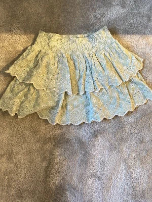 Grön volangkjol från Lindex - Supersöt grön volangkjol från Lindex med broderade blommor. Kjolen har smock i midjan och flera lager med vågig kant. Storlek 146/152 men kan användas upp till 158 eller 164