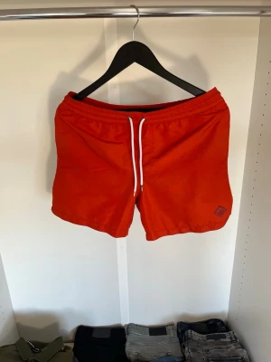Röda badbyxor från J.Lindeberg - Snygga röda badbyxor från J.Lindeberg med vitt dragsnöre i midjan och en liten logotyp på vänster ben. Klassisk modell med elastisk midja och snabbtorkande material. Hör gärna av dig vid frågor!