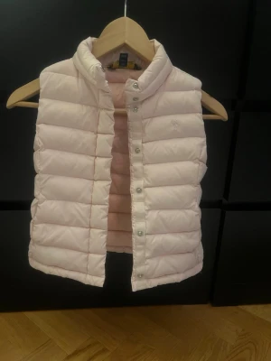 Ljusrosa dunväst från Ralph Laurent  - Supersnygg ljusrosa dunväst från Ralph Laurent med hög krage och quiltad design. Västen har tryckknappar framtill och diskret logga broderad på bröstet. Perfekt att slänga över en hoodie eller tröja för extra värme och stil.