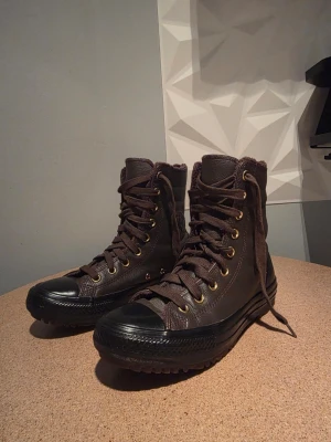 Bruna höga kängor från Converse - Snygga bruna höga kängor från Converse med klassisk stjärnlogga på sidan. Kängorna har grov sula, guldiga öljetter och är gjorda i läder med textildetaljer. Perfekta för dig som vill ha en cool och robust stil.