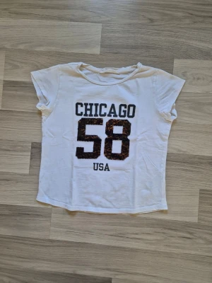 Vit t-shirt med CHICAGO 58-paljetter - Vit t-shirt med korta ärmar och tryck framtill med texten 'CHICAGO 58 USA'. Siffran 58 är vändbara paljetter för en cool kontrast. T-shirten har en avslappnad passform och är gjord i mjuk bomull, perfekt till vardagslooken.