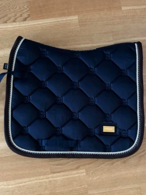 Marinblått schabrak från Equestrian Stockholm - Säljer ett marinblått schabrak från Equestrian Stockholm med quiltat mönster och dekorativ kant i beige och vitt. Schabraket har en guldfärgad logoplatta och är tillverkat i glansigt material. Perfekt för dig som vill ha stil på ridturen. Knappt andvända.