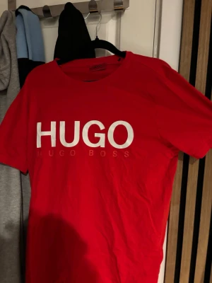 Röd HUGO BOSS t-shirt med tryck - Riktigt snygg röd t-shirt från HUGO BOSS med stort vitt HUGO-tryck på bröstet. Klassisk passform och rund halsringning, perfekt för en stilren och modern look. Tillverkad i mjuk bomull som känns skön mot huden.