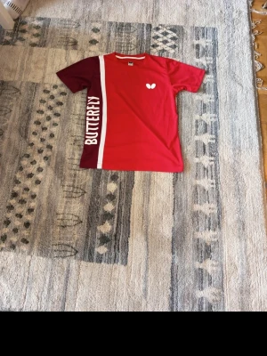 Röd sporttröja från Butterfly - Röd och vinröd sporttröja från Butterfly med vit logga och text på framsidan. Tröjan har en vit vertikal linje och är tillverkad i ett lätt, ventilerande material som passar perfekt för intensiva sportaktiviteter. Unisex-modell med kort ärm.