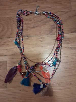 Färgglatt halsband med pärlor och tofsar - Unikt halsband med flera rader små färgglada pärlor, dekorerat med tofsar i blått, beige och lila samt fjädrar och små berlocker. Halsbandet har inslag av guldtonade kedjor och detaljer i plast och metall. Perfekt för dig som gillar boho-stil och färg.