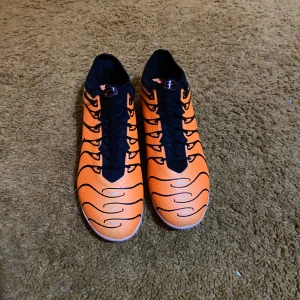 Orange fotbollsskor från Nike - Säljer ett par grymma orangea fotbollsskor från Nike i nytt skick. De har en snygg svart snörning och en cool design med svarta detaljer. Perfekta för fotbollssäsongen och ger bra grepp på planen. Skorna är lätta och bekväma, redo för match!