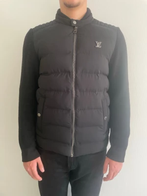 Svart dunväst från Louis Vuitton - Snygg svart dunväst från Louis Vuitton med diskret LV-logga på bröstet. Västen har quiltad design, hög krage och dragkedja framtill samt två fickor med tryckknappar. Perfekt för lager-på-lager och ger en clean look.