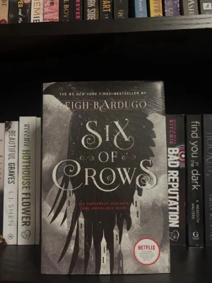 Six of Crows av Leigh Bardugo - En spännande fantasybok med svart och grått omslag, prydd med en stor korp och gotiska byggnader. Boken är på engelska och har en Netflix-sticker på framsidan. Perfekt för dig som gillar äventyr och magi.
