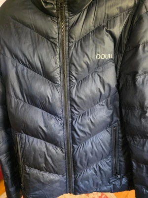 Svart dunjacka från Hugo boss - Snygg svart dunjacka från Liu Jo med dragkedja framtill och två sidofickor med dragkedja. Jackan har ett stilrent mönster i chevron-stil och diskret logga på bröstet. Perfekt för kalla vinterdagar och riktigt skön att bära.