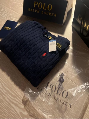 Mörkblå kabelstickad tröja Polo Ralph Lauren - Mörkblå kabelstickad tröja från Polo Ralph Lauren med rund halsringning och klassisk röd broderad logga på bröstet. Tröjan är i mjukt material och har ribbade muddar vid ärmslut och nederkant. Perfekt för en stilren och tidlös look.tilbehöret ingår inte men tagen som sitter fast ingår.Stolek=Si herr storlek.
