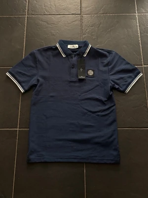 Marinblå pikétröja från Stone Island - Snygg marinblå pikétröja från Stone Island med vitrandiga detaljer på krage och ärmslut. Tröjan har klassisk krage, knappar framtill och ikonisk logotyp på bröstet. Perfekt för en stilren och sportig look.