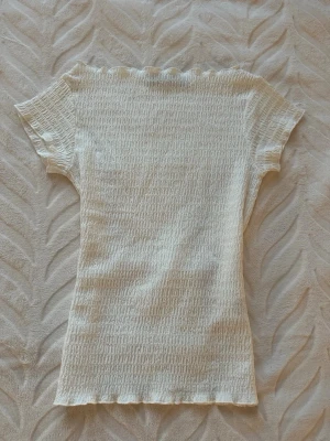 Vit ribbad topp med knyt i midjan - Superfin vit ribbad topp med korta ärmar och knytband i midjan. Toppen har en lätt vågig kant vid hals och ärmslut, och är tillverkad i ett stretchigt material som sitter snyggt på kroppen. Perfekt att styla med jeans eller kjol. Den är knappt använd och har inga fräckar ser nästan helt ny ut pris kan diskuteras 
