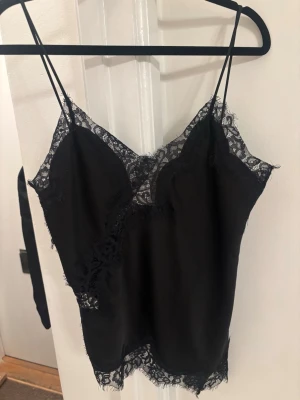 Ny Zara topp Svart spetslinnetopp med smala band - Snygg svart linnetopp med tunna axelband och dekorativa spetsdetaljer längs urringning och nederkant. Asymmetrisk kant ger en unik look och materialet känns lent och följsamt. Perfekt för dig som vill ha en trendig och elegant topp. Ny från Zara 