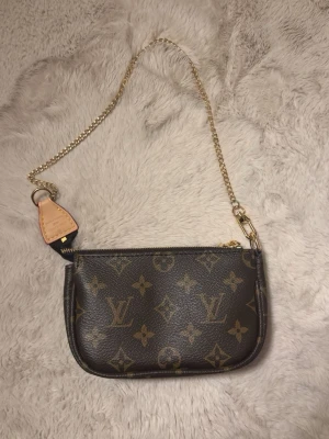 Brun väska - Snygg liten handväska i klassiskt brunt monogrammönster. Väskan har guldfärgad kedja som axelrem och detaljer i läder. Perfekt för att bära det viktigaste och ger en lyxig touch till din outfit.