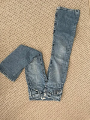 Blå bootcut jeans med slitningar - Säljer ett par ljusblå bootcut jeans med snygga slitningar och klassisk femficksmodell från Gina tricot. Jeansen har normal midja och dragkedja med knapp. Perfekta för en avslappnad och trendig look.